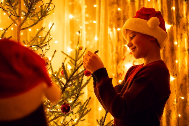 Glimlachend jongetje in rode trui en kerstmuts opknoping kerstbal aan de kerstboom