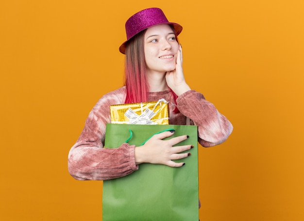 Glimlachend kijkende jonge, mooie meid met een feesthoed met een cadeauzakje en zet de hand op de wang geïsoleerd op een oranje muur