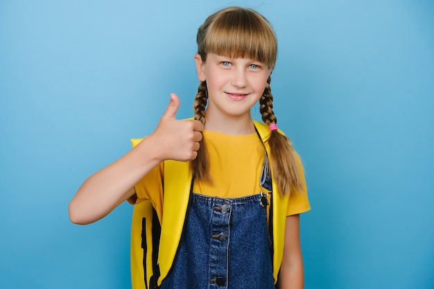 Glimlachend kind basisschoolmeisje met gele rugzak duim opdagen, geïsoleerd over blauwe kleur achtergrond in studio. Gelukkige blonde kindstudent die vrijheid viert en de beste onderwijskeuze aanbeveelt