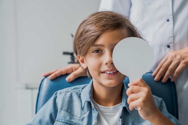 Glimlachend kind bedekt het oog met een medisch hulpmiddel voor het controleren van de gezichtsscherpte in de medische kliniekkast