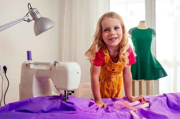 Glimlachend klein blond meisje aan tafel met naaimachine met oefenpop