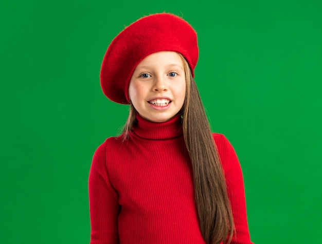 Glimlachend klein blond meisje met een rode baret kijkend naar de voorkant geïsoleerd op een groene muur met kopieerruimte