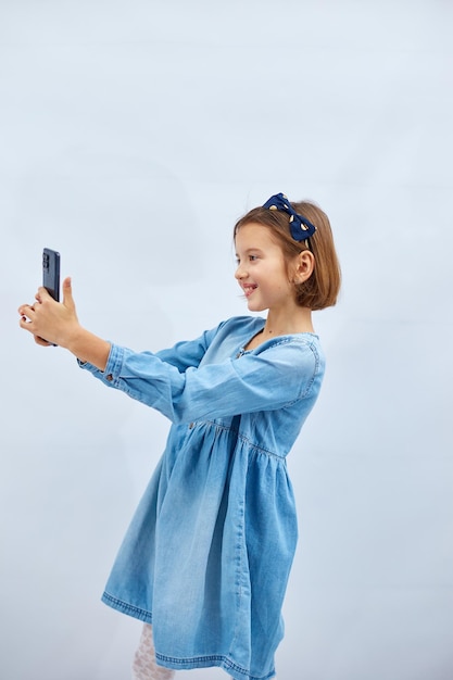 Glimlachend klein meisje in casual denim jurk houdt smartphone selfie