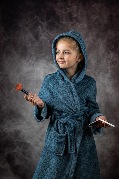Glimlachend klein meisje in een badjas en borstel geïsoleerd op grijze achtergrond Makeup Model test