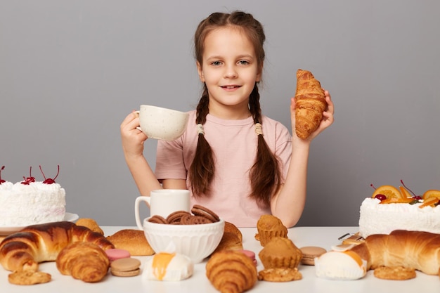 Glimlachend klein meisje met staartjes zittend aan tafel geïsoleerd over de grijze achtergrond genietend van verse smaak gebakken croissant thee drinken camera kijkend