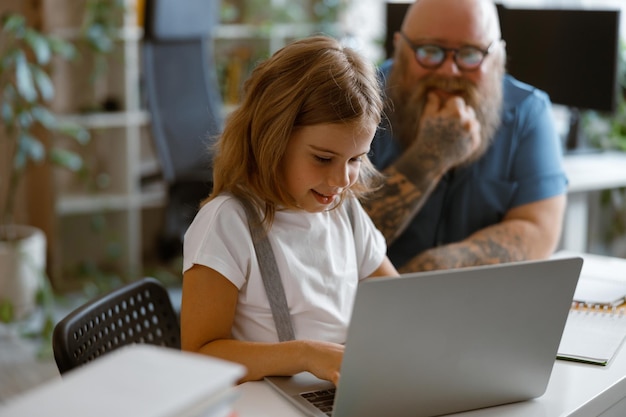 Glimlachend klein meisje typt op laptop terwijl vader in de buurt aan tafel zit in een lichte kamer