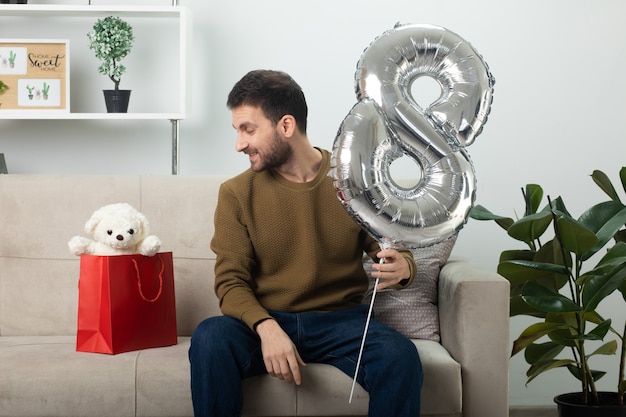 Glimlachend knappe man met ballon vormige acht en kijken naar teddybeer in cadeauzakje zittend op de bank in de woonkamer op maart internationale Vrouwendag