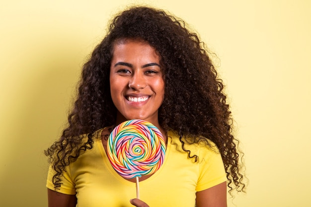 Glimlachend krullend afrikaans meisje in helder geel t-shirt met snoep lolly. jonge vrolijke vrouw die voorzijde met lolly verbergt