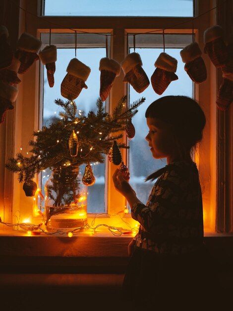 Glimlachend lief kindmeisje dat met Kerstmis bij het raam staat Met kleurrijke lampjes van de kerstboom op de achtergrond, selectieve aandacht.