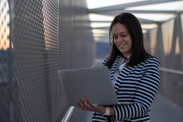 Glimlachend meisje dat met behulp van laptop op een brug staat Glimlachende vrouw die met behulp van laptop op de gang van een brug staat Concept vrouwelijke ondernemer die laptop op een brug gebruikt