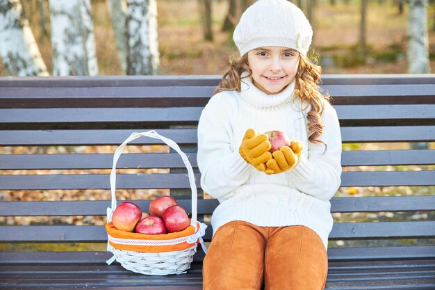 Glimlachend meisje in een witte trui en baret zit in de herfst op een bankje in het park met een mand met appels. hoge kwaliteit foto