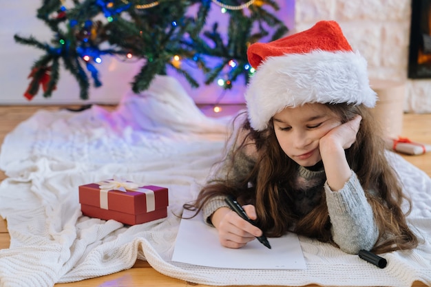 Glimlachend meisje in kerstmuts schrijven brief voor geschenken aan de kerstman.