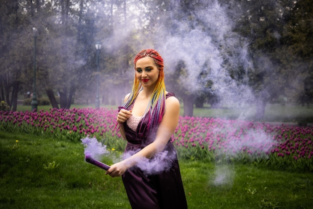 Glimlachend meisje in paarse satijnen jurk met lange veelkleurige vlechten en opvallende glitter make-up. ongewone kunstmatige rook van paarse kleur bedekt het meisje in het lentepark.