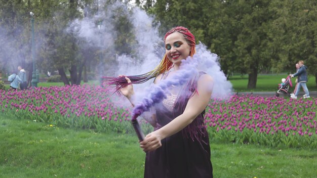 Glimlachend meisje in paarse satijnen jurk met lange veelkleurige vlechten en opvallende glitter make-up. Rook van paarse kleur bedekt het meisje in het lentepark