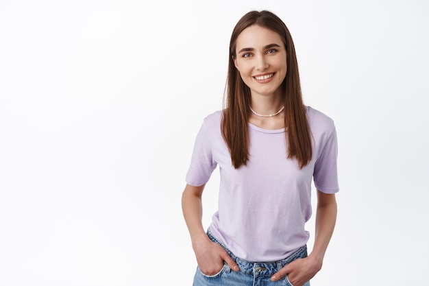 Foto glimlachend meisje in t-shirt ziet er vriendelijk en gelukkig uit voor de camera, ontspannen pose van een vrouw die tegen een witte achtergrond staat met de handen in de zakken.