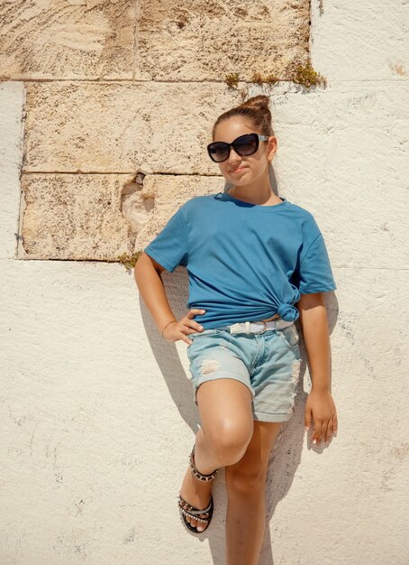 Glimlachend meisje met een t-shirt, korte broek en zonnebril die in de buurt van een stenen muur buiten blijft en recht in de camera kijkt. t-shirtmodel