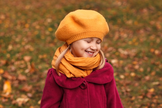 Glimlachend meisje met gesloten ogen in een gele baret en herfstkleren op een herfstachtergrond