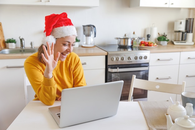 Glimlachend meisje met kerstmuts zwaaiende hand video bellen familie door webcam. Vrouw met laptop met virtuele vergadering chat op vakantie zittend op de keuken thuis. Fijne kerst en nieuwjaar nieuw normaal