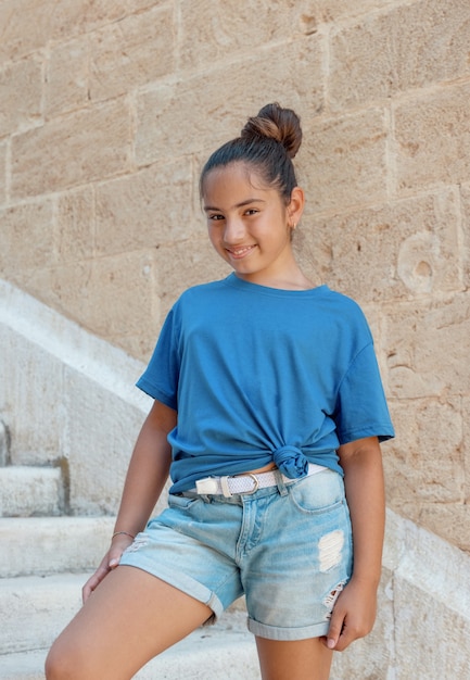Glimlachend meisje met t-shirt en korte broek die buiten op stenen trappen blijft, glimlachend en recht in de camera kijkt