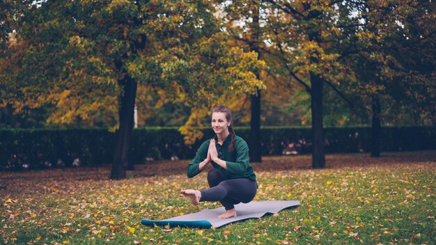 Foto glimlachend meisje professionele yoga-instructeur doet evenwichtsoefeningen staande op één been op mat op gras in park mooie herfst natuur is zichtbaar