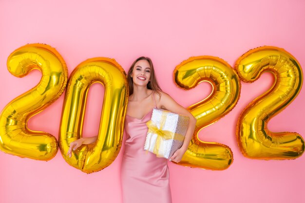 Glimlachend meisje staat in de buurt van luchtballonnen houdt geschenkdoos op roze achtergrond nieuwjaarsviering