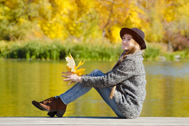 Glimlachend meisje zittend op de pier met herfstbladeren in handen. Zonnige dag