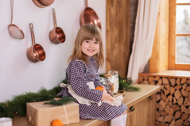 Glimlachend meisje zittend op houten tafelblad op keuken met kerstcadeaus en mandarijnen