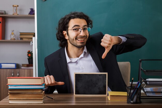 Glimlachend met duim omlaag mannelijke leraar die een bril draagt met een mini schoolbord aan tafel met schoolhulpmiddelen in de klas