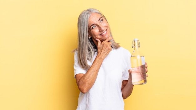 Glimlachend met een gelukkige, zelfverzekerde uitdrukking met de hand op de kin, zich afvragend en opzij kijkend en een waterfles vasthoudend