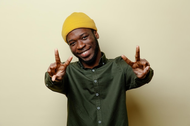 Glimlachend met vredesgebaar jonge Afro-Amerikaanse man in hoed met groen shirt geïsoleerd op een witte achtergrond