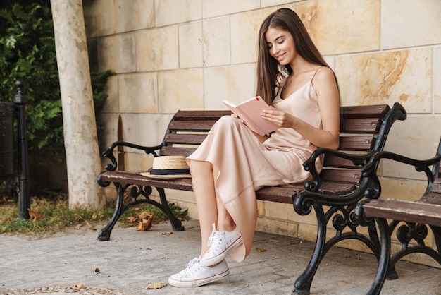 Glimlachend mooi jong meisje in zomerjurk zittend op een bankje buiten, een boek lezend