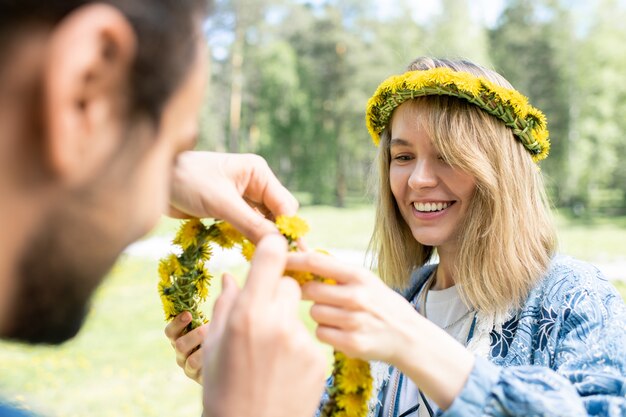 Glimlachend mooi meisje bijstaan vriendje terwijl ze paardebloem krans buitenshuis maken