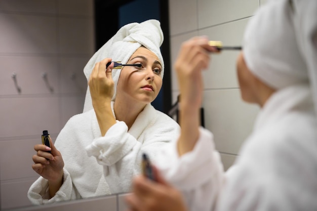 Glimlachend mooi meisje in een badjas die make-up aanbrengt bij de spiegel