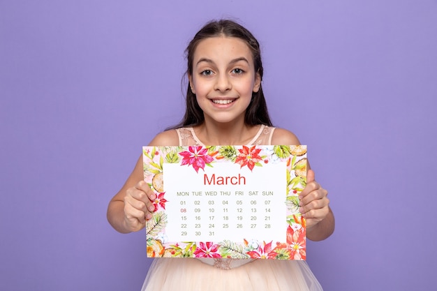 Glimlachend mooi meisje op de dag van de gelukkige vrouw met kalender geïsoleerd op blauwe muur