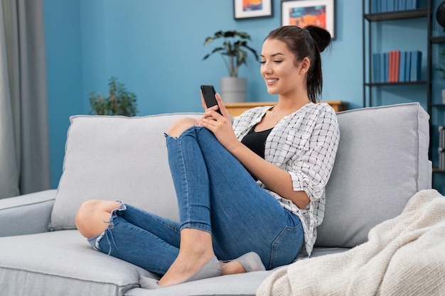 Glimlachend mooi tienermeisje zit op de bank in de woonkamer met telefoon