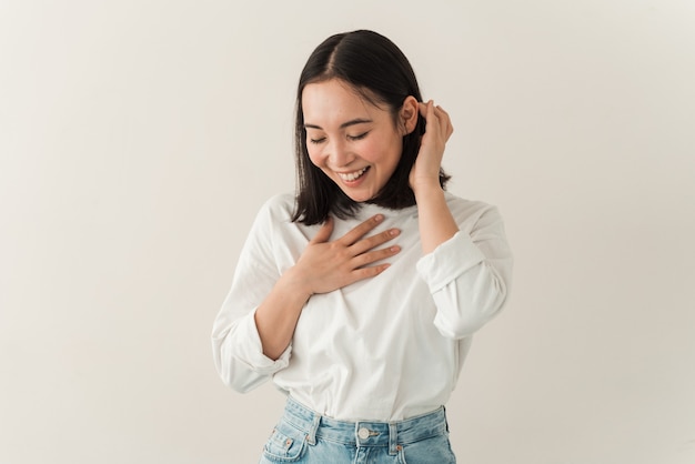 Glimlachend, mooie Aziatische brunette poseren op een witte muur achtergrond. Jonge vrouw schaamt zich terwijl ze in de studio staat met een blinde muur