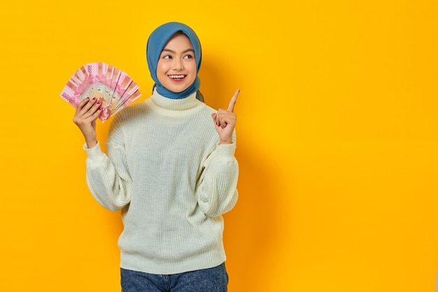 Glimlachend mooie Aziatische moslimvrouw in witte trui met contant geld in Indonesische rupiah bankbiljetten en wijzende vinger opzij geïsoleerd over gele achtergrond Mensen religieuze levensstijl concept
