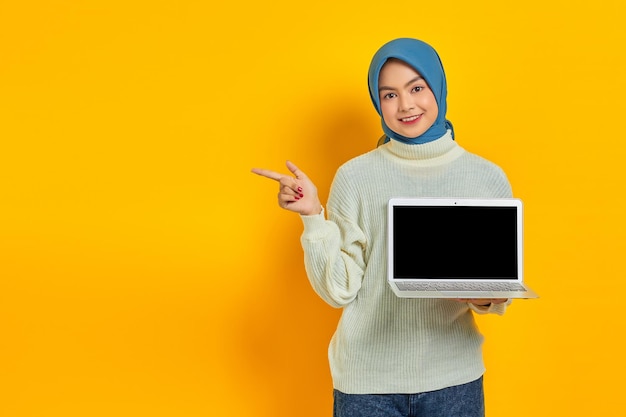 Glimlachend mooie Aziatische vrouw in witte trui en hijab met een laptop met leeg scherm en wijzende vingers op kopie ruimte geïsoleerd over gele achtergrond mensen religieuze levensstijl concept