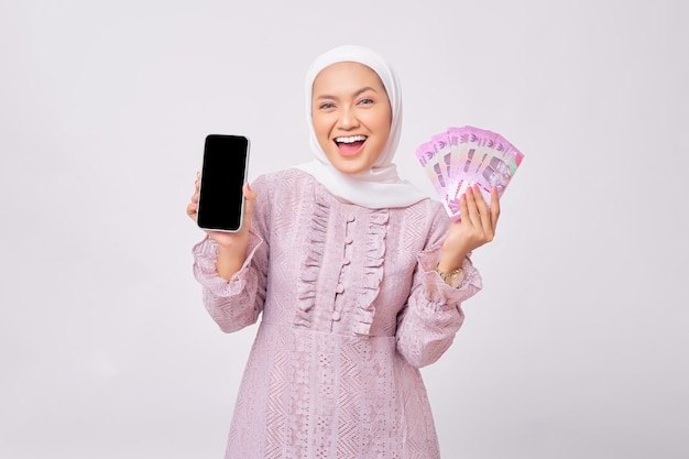 Glimlachend mooie jonge Aziatische moslimvrouw dragen hijab en paarse jurk tonen mobiele telefoon met leeg scherm en contant geld rupiah bankbiljetten geïsoleerd op witte studio achtergrond