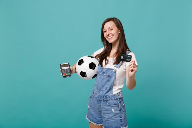 Glimlachend mooie jonge vrouw voetbalfan ondersteuning favoriete team met voetbal, draadloze moderne bankbetaalterminal te verwerken, creditcardbetalingen geïsoleerd op blauwe turkooizen achtergrond te verwerven.