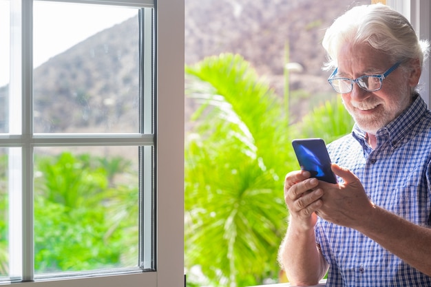 Glimlachend mooie senior man bij het raam met behulp van mobiele telefoon