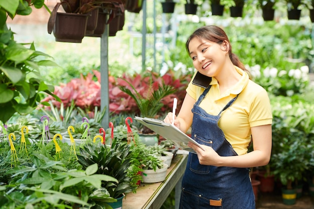 Glimlachend mooie tuincentrummedewerker die telefonisch met de klant praat en aantekeningen maakt in document