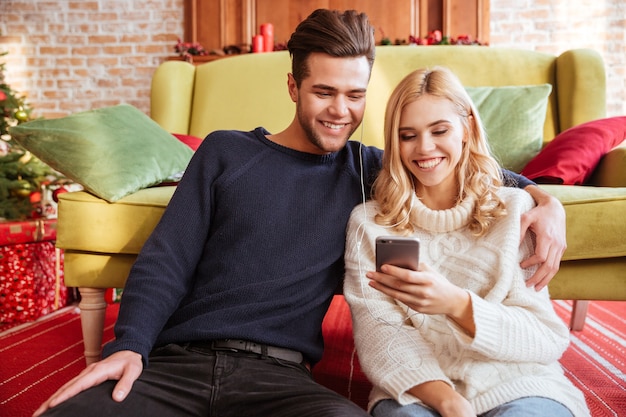 Glimlachend paar in truien die bij de bank zitten en naar de telefoon kijken