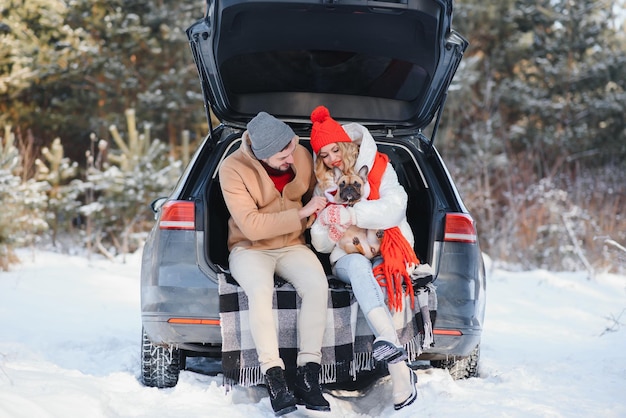 Glimlachend paar met hond zittend in open suv-kofferbak in besneeuwd bos genieten van elkaar in actieve wintervakanties