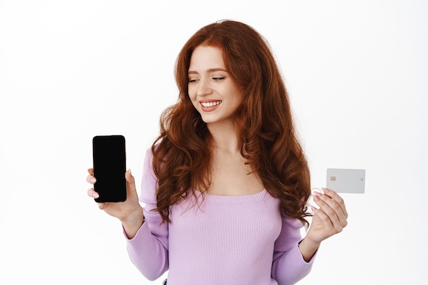 Glimlachend roodharig vrouwelijk model met creditcard, kijkend naar een leeg smartphonescherm, app voor mobiele telefoons aanbevelend, staande op wit