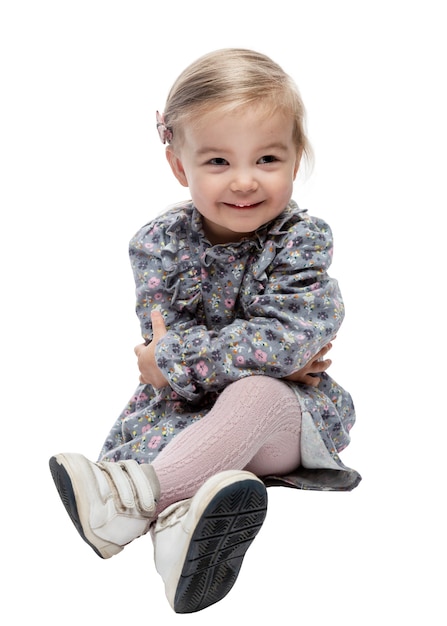 Glimlachend schattig klein meisje van 2 jaar oud in een jurk zit met gekruiste benen op de vloer. geïsoleerd op een witte achtergrond. verticaal.
