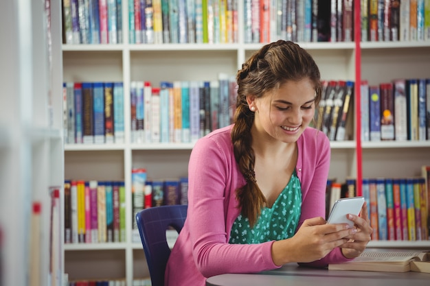 Glimlachend schoolmeisje die mobiele telefoon in bibliotheek met behulp van