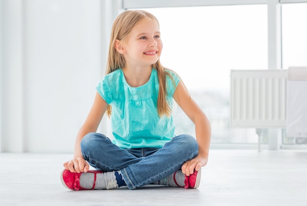 Glimlachend schoolmeisje op vloer dichtbij venster binnenshuis.