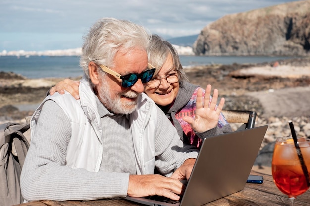 Foto glimlachend senior paar in videochat met laptop genieten van zonnige dag buiten op het strand zittend aan de bar