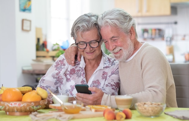 Glimlachend senior paar op zoek naar mobiel tijdens het ontbijt thuis